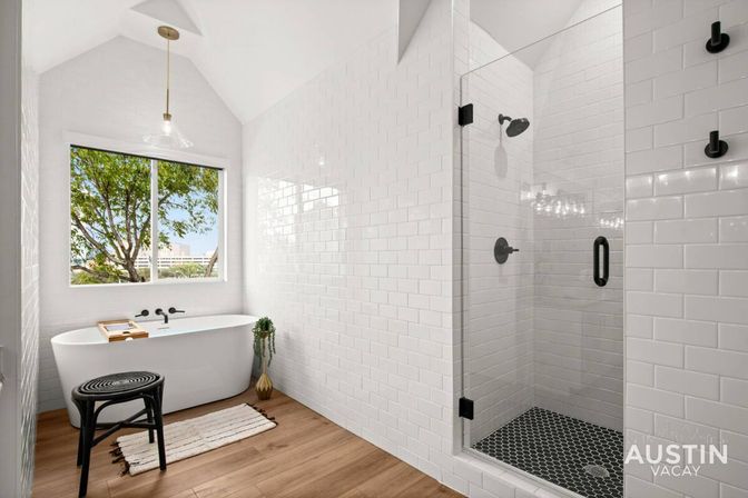 Bright modern bathroom with white subway tile, freestanding tub beneath a tree-view window, black matte fixtures, glass-enclosed shower with dark hex-tile floor, wood-look flooring, pendant light, stool and bath tray.