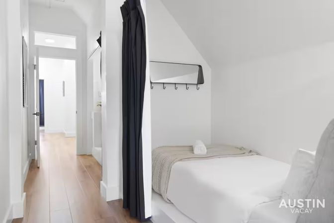 Bright minimalist apartment hallway with warm wood floors leading to a cozy bed tucked into a white alcove with a black privacy curtain, wall mirror and hooks.