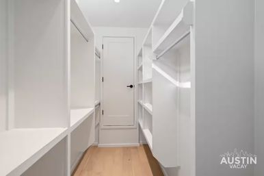 Sleek bright minimalist walk-in closet with white built-in shelves and hanging rods, light hardwood floor and a closed door at the end.