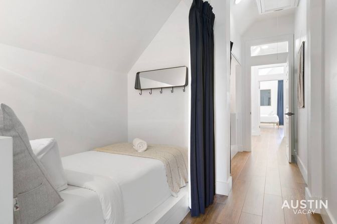 Bright minimalist bedroom nook with a single bed, rolled towel, wall mirror with hooks and navy privacy curtain opening onto a sunny hallway with wood floors.