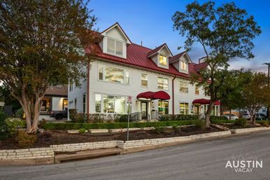 Dusk view of a white three-story building with a red metal roof, dormer windows and red awnings, framed by trees along a quiet Austin street