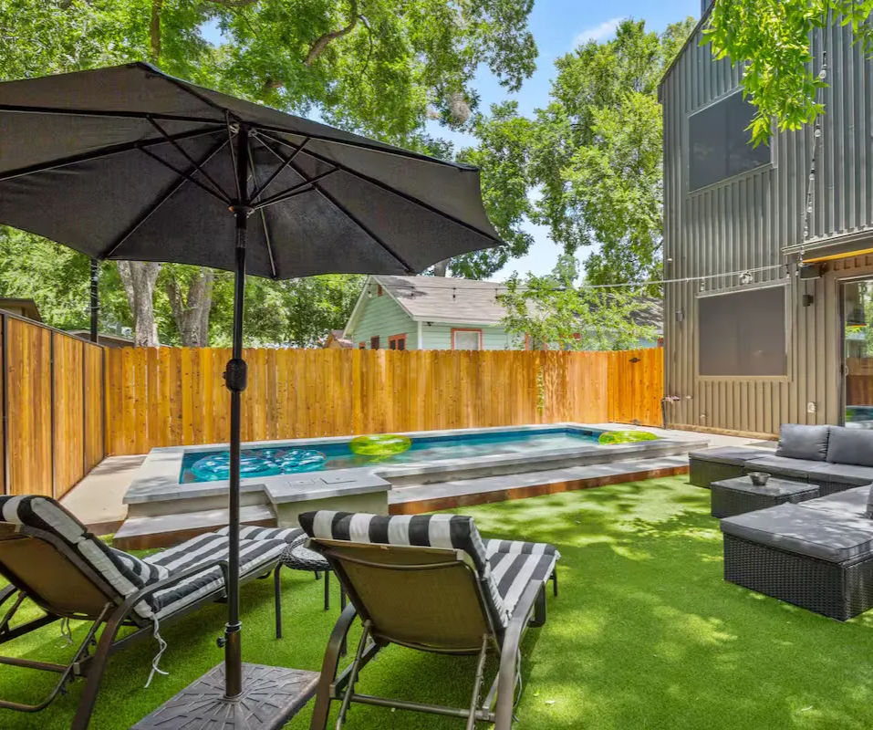 Sunny modern backyard with narrow plunge pool on a raised concrete deck, artificial turf lawn, black patio umbrella, striped chaise lounges and gray wicker sectional beside a wooden privacy fence and leafy trees