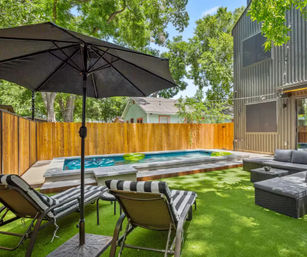 Sunny modern backyard with narrow plunge pool on a raised concrete deck, artificial turf lawn, black patio umbrella, striped chaise lounges and gray wicker sectional beside a wooden privacy fence and leafy trees