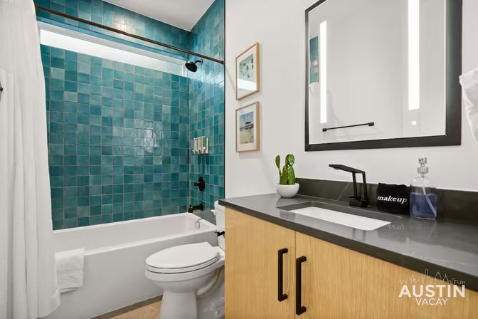 Bright modern bathroom with teal square-tile shower surround, white tub and toilet, wood vanity with dark countertop and black faucet, illuminated mirror and small potted plant — Austin