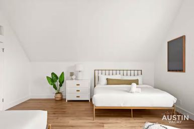 Minimalist Austin bedroom with brass-framed bed, white linens and olive lumbar pillow, white nightstand with lamp, potted green plant and warm wood floors.