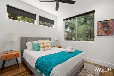Bright modern bedroom with gray upholstered bed, teal throw and pillows, wooden mid-century nightstands, ceiling fan and large windows looking out to leafy trees in Austin, TX