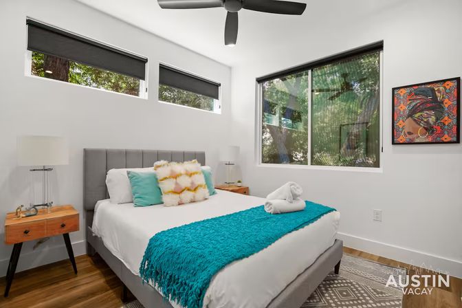 Bright modern bedroom with gray upholstered bed, teal throw and pillows, wooden mid-century nightstands, ceiling fan and large windows looking out to leafy trees in Austin, TX
