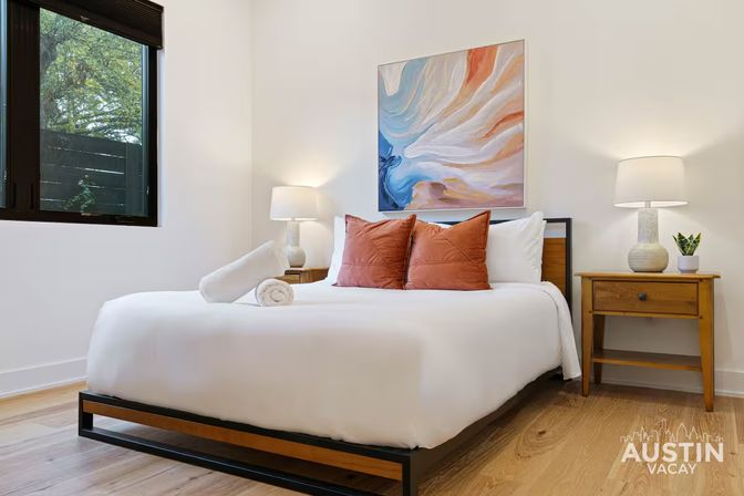 Bright, airy vacation rental bedroom with crisp white linens, two terracotta throw pillows and a rolled towel on a low wood-and-metal platform bed, abstract pastel wall art, matching wooden nightstands with table lamps and a small plant, and a window showing greenery.