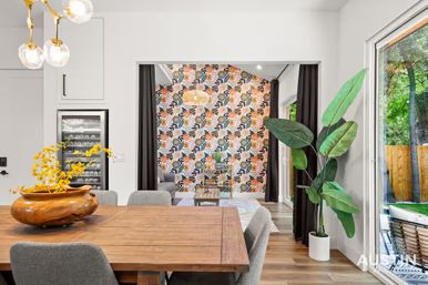 Bright modern open-plan dining room with a wooden table and gray upholstered chairs, a large tropical potted plant by sliding glass doors to a backyard patio, and a vibrant floral accent wall in the adjoining living area.