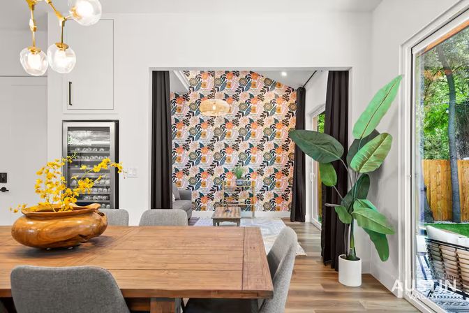 Bright modern open-plan dining room with a wooden table and gray upholstered chairs, a large tropical potted plant by sliding glass doors to a backyard patio, and a vibrant floral accent wall in the adjoining living area.