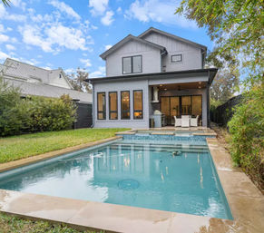 Modern gray two-story suburban home with a rectangular backyard swimming pool, covered patio with outdoor dining and grill, lawn and privacy hedges under a blue sky