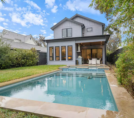 Modern gray two-story suburban home with a rectangular backyard swimming pool, covered patio with outdoor dining and grill, lawn and privacy hedges under a blue sky