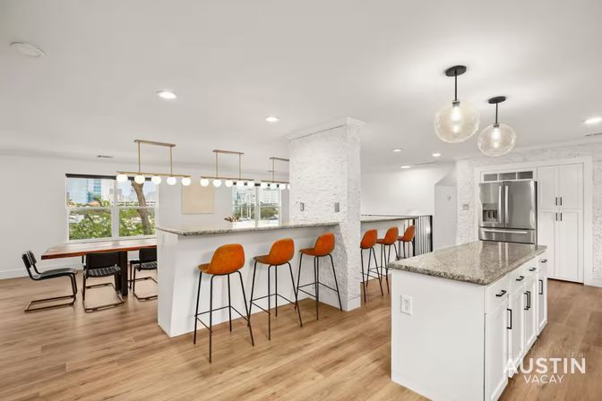 Bright modern open-concept Austin kitchen and dining area with white cabinets, granite island, breakfast bar with orange leather stools, globe pendant lights, hardwood floors and city-view windows.