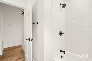 Bright modern bathroom with white subway-tiled tub/shower and matte-black fixtures, open door to hardwood hallway with a playful animal wall mount peeking