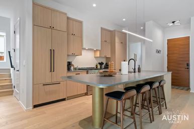 Bright modern Austin vacation-rental kitchen with light wood cabinets, integrated appliances, large island with four bar stools, black faucet and linear pendant lighting.