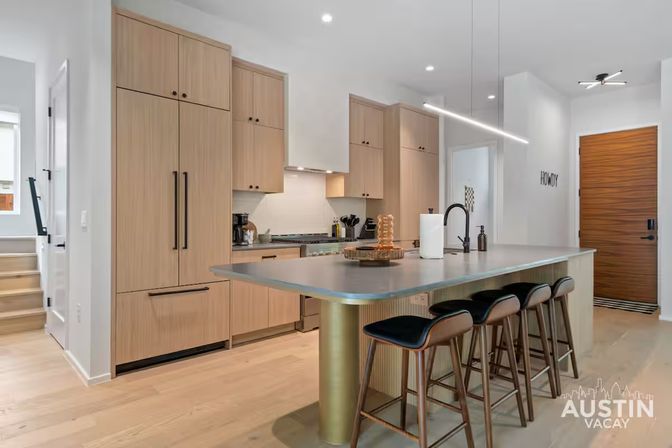 Bright modern Austin vacation-rental kitchen with light wood cabinets, integrated appliances, large island with four bar stools, black faucet and linear pendant lighting.