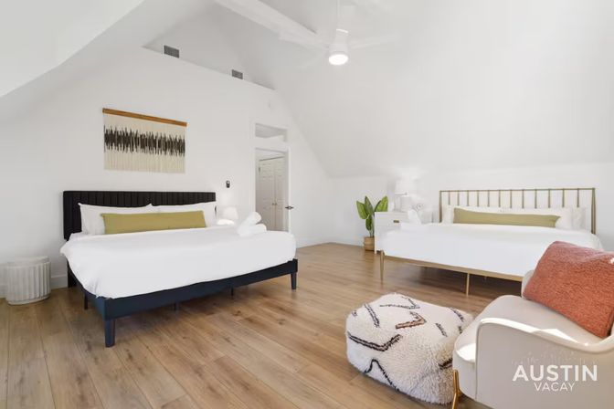 Bright airy attic-style vacation rental bedroom with two beds, vaulted white ceiling, wood floors, ceiling fan, potted plant, cozy chair and fluffy pouf