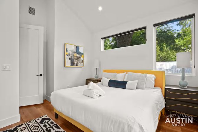 Bright modern Austin vacation rental bedroom with a yellow-upholstered bed, crisp white linens, wood floors, abstract wall art and tree-lined windows.