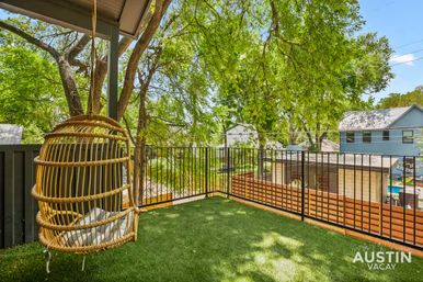 Sunny Austin balcony with a hanging rattan egg chair, artificial turf flooring, black metal railing, and leafy trees overlooking neighboring houses.