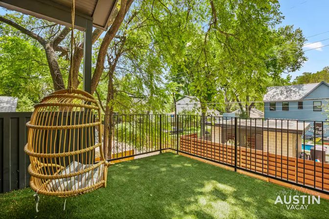 Sunny Austin balcony with a hanging rattan egg chair, artificial turf flooring, black metal railing, and leafy trees overlooking neighboring houses.