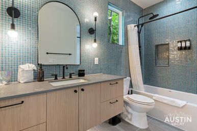 Bright modern Austin bathroom with blue mosaic tile walls, light wood vanity and arched mirror, matte-black faucet and sconces, tub-shower with white curtain and small window showing trees.