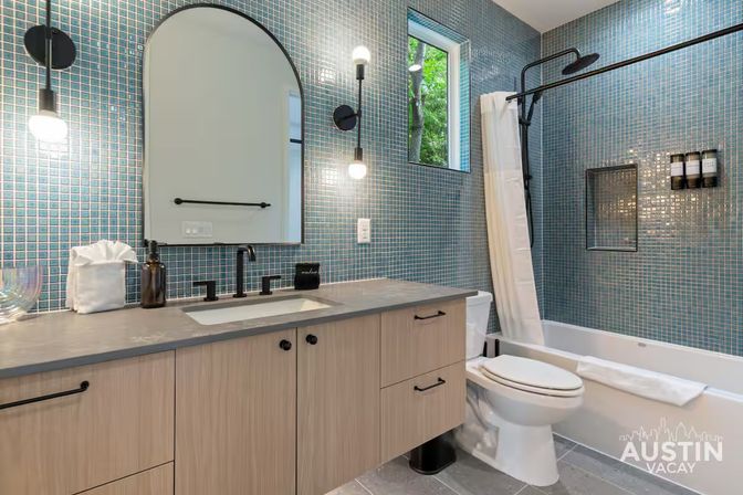 Bright modern Austin bathroom with blue mosaic tile walls, light wood vanity and arched mirror, matte-black faucet and sconces, tub-shower with white curtain and small window showing trees.