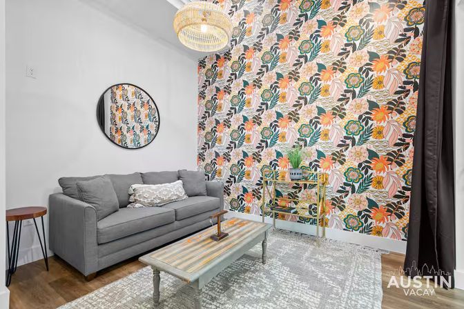 Cozy modern living room with bold coral-and-green floral accent wall, gray sofa with decorative pillow, round wall mirror, striped wood coffee table, rattan pendant light, bar cart and textured area rug.