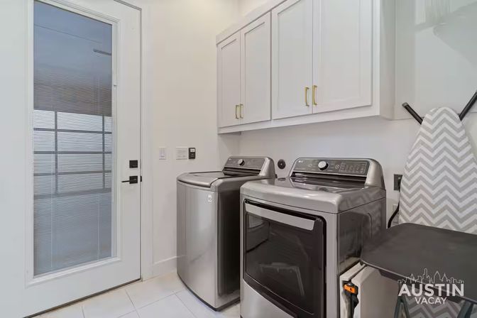 Bright modern laundry room with sleek stainless-steel top-load washer and front-load dryer, white upper cabinets with brass handles, chevron ironing board, tiled floor and frosted-glass door.