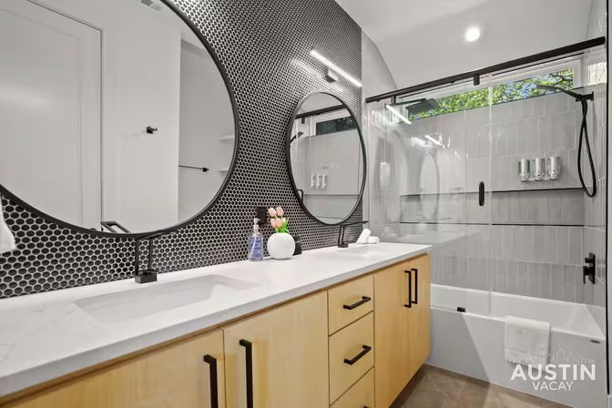 Sleek modern bathroom with double sinks and round mirrors, white quartz countertop, light wood cabinets with black handles, black penny-tile backsplash and matte-black faucets, glass sliding shower enclosure with tub and mounted dispensers and a high window showing greenery.