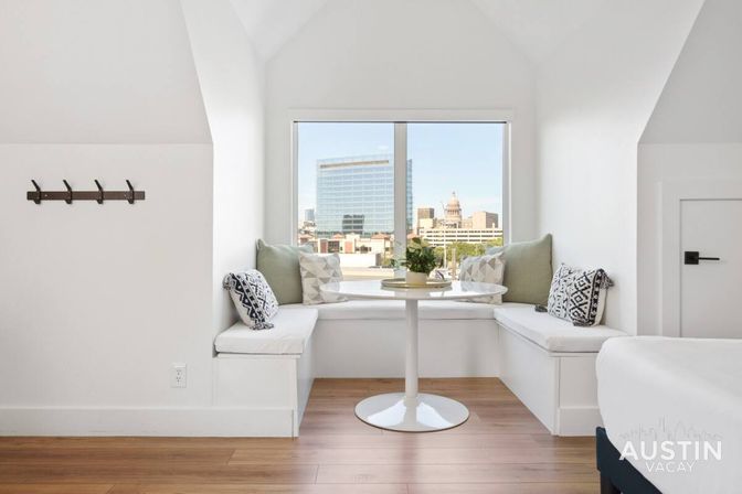 Bright attic window nook with built-in bench seating, round pedestal table, patterned pillows, plant, and Austin skyline view.