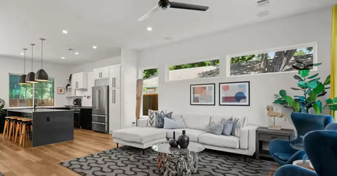 Sunlit modern open-plan living room and kitchen — white L-shaped sectional, driftwood coffee table, geometric rug, blue accent chairs, black kitchen island with wooden stools and pendant lights, hardwood floors, clerestory windows and indoor plants.