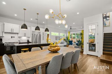 Bright, modern open-concept kitchen and dining area with a rustic wooden table and gray upholstered chairs, black island with yellow stools and pendant lights, brass orb chandelier, stainless steel appliances, and a cozy living space with blue accent chairs and sliding glass doors to the backyard.