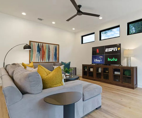 Bright modern living room with light-gray sectional sofa and mustard throw pillows, abstract wall art, arc floor lamp and ceiling fan, hardwood floors and a wooden media console with TV showing streaming apps.