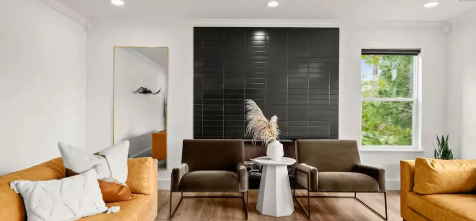 Modern living room with mustard sofas, two brown accent chairs flanking a white pedestal table topped with pampas grass, black tile feature wall, full-length mirror, window showing green trees, and warm wood floors.