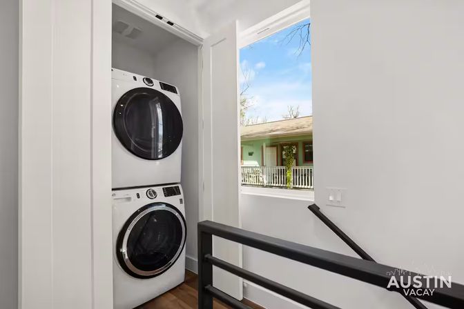 Cozy upstairs laundry nook with stacked front-load washer and dryer in a white closet, natural light from a window overlooking a neighboring porch and modern black stair railing.
