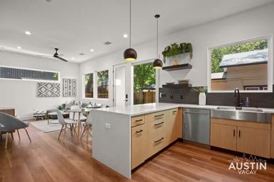 Bright modern open-concept kitchen and living room with white island and light wood cabinets, stainless steel sink and dishwasher, black pendant lights, hardwood floors, dining table and cozy seating, large windows revealing a leafy backyard.