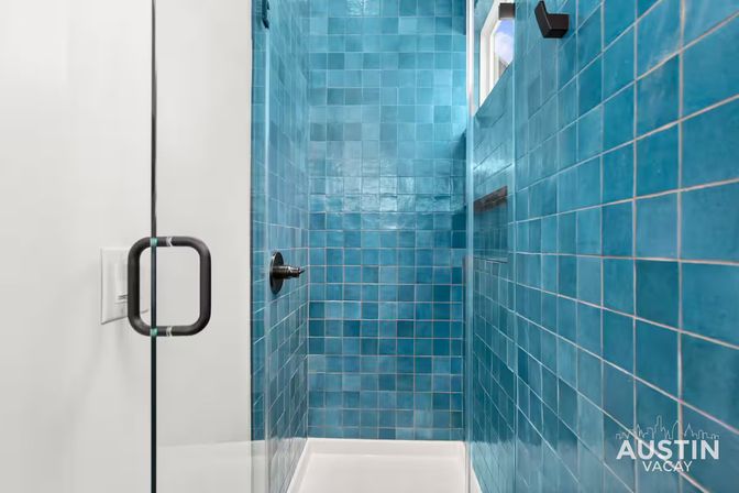 Vibrant turquoise-blue tiled walk-in shower with glossy square tiles, frameless glass door and black handle, built-in niche and dark shower control.