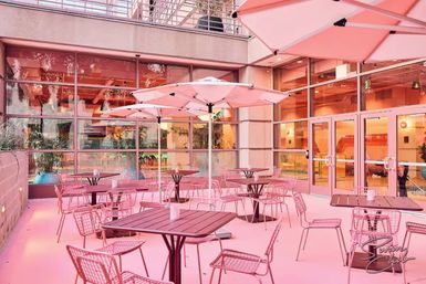 Vibrant pink urban patio with alfresco seating — umbrella-shaded tables and wire metal chairs beside a glass-front building