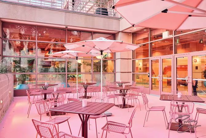 Vibrant pink urban patio with alfresco seating — umbrella-shaded tables and wire metal chairs beside a glass-front building