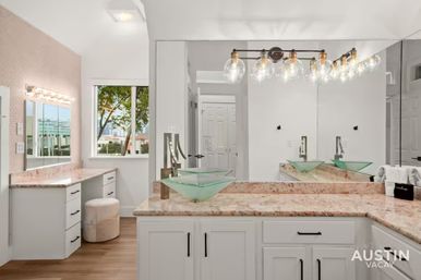 Bright modern vacation-rental bathroom in Austin with twin green glass vessel sinks on marble countertops, white shaker cabinets, Hollywood-style vanity lights, large wall mirror, window with tree and city view, and wood-look flooring.