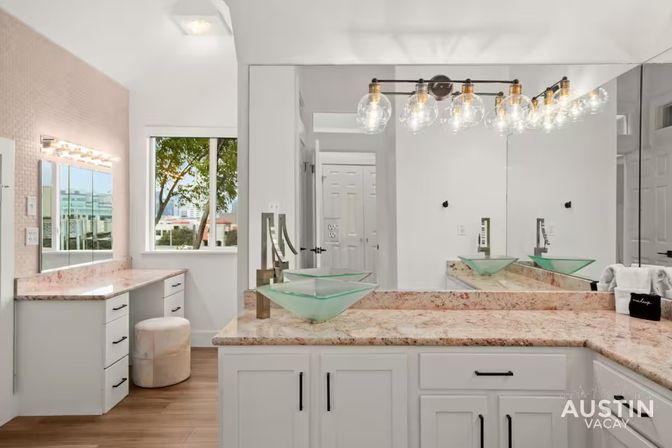 Bright modern vacation-rental bathroom in Austin with twin green glass vessel sinks on marble countertops, white shaker cabinets, Hollywood-style vanity lights, large wall mirror, window with tree and city view, and wood-look flooring.