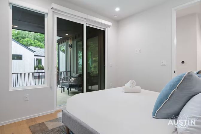 Bright modern bedroom in Austin with sliding glass doors to a covered balcony featuring wicker chairs, crisp white bedding, blue accent pillows, and hardwood floors.