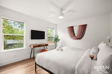 Bright modern Austin bedroom with large windows looking out to green trees, wall-mounted TV above a mid-century wooden console, potted plant, cozy king bed with crisp white linens and pillows, ceiling fan, and colorful woven wall hanging.
