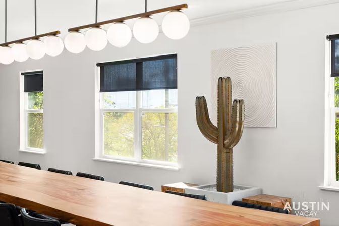 Bright modern Austin conference room with a long wooden table, globe pendant lights, three windows with dark roller shades, and a metal saguaro cactus sculpture against a white wall.