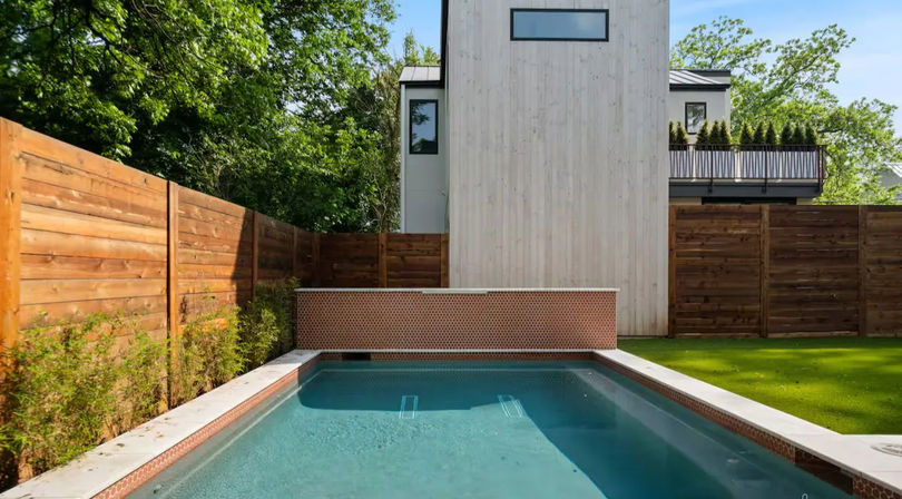 Sunlit modern residential backyard with a narrow rectangular swimming pool and clear blue water, flanked by wooden privacy fences, a raised mosaic-tiled end wall, contemporary white-clad house facade and green lawn.