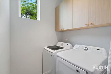 Bright minimalist laundry room with matching white washer and dryer, light wood wall cabinets, and a small high window revealing green tree branches.