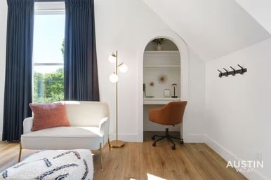 Sunlit modern living nook with a white loveseat and pink throw pillow by tall dark curtains, brass floor lamp on hardwood floors, arched built-in desk with a brown swivel chair, and wall hooks.