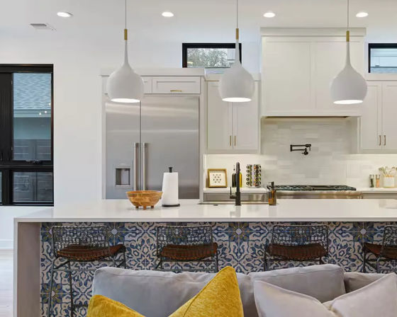 Bright modern open-concept kitchen with white waterfall island, patterned blue-and-white tile bar front, three white pendant lights, stainless steel fridge and gas range, wire bar stools, and gray sofa with a yellow accent pillow in the foreground.