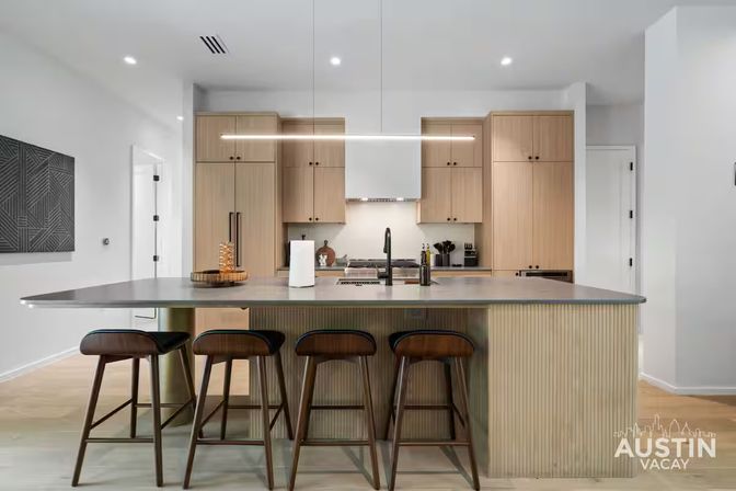 Bright modern kitchen with a large island and four wooden bar stools, light wood cabinets, integrated sink, stainless appliances and a sleek linear pendant light.