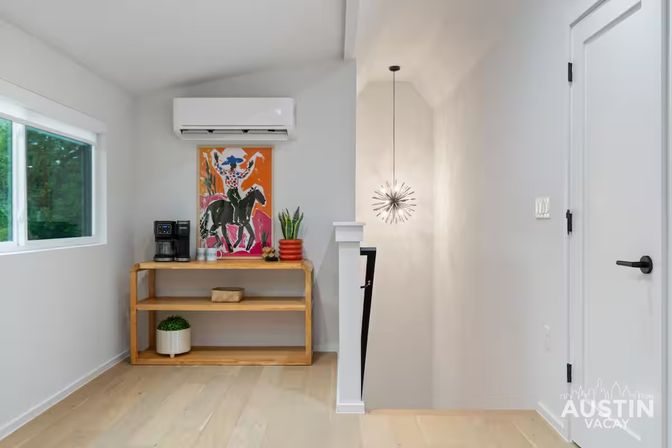 Bright upstairs landing in a modern home with a wall-mounted air conditioner above a wooden console used as a mini coffee station, colorful cowboy print, potted plants, starburst pendant light, window with greenery, and white door by the stairs.